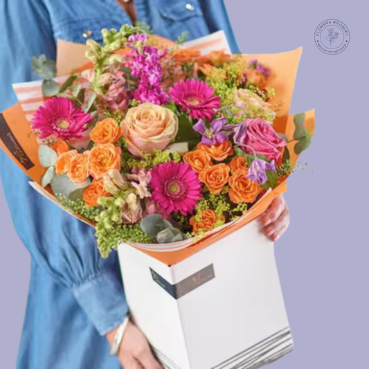 Bright orange and pink flower bouquet featuring roses, gerbera daisies, and lavender stock in a premium white gift box.